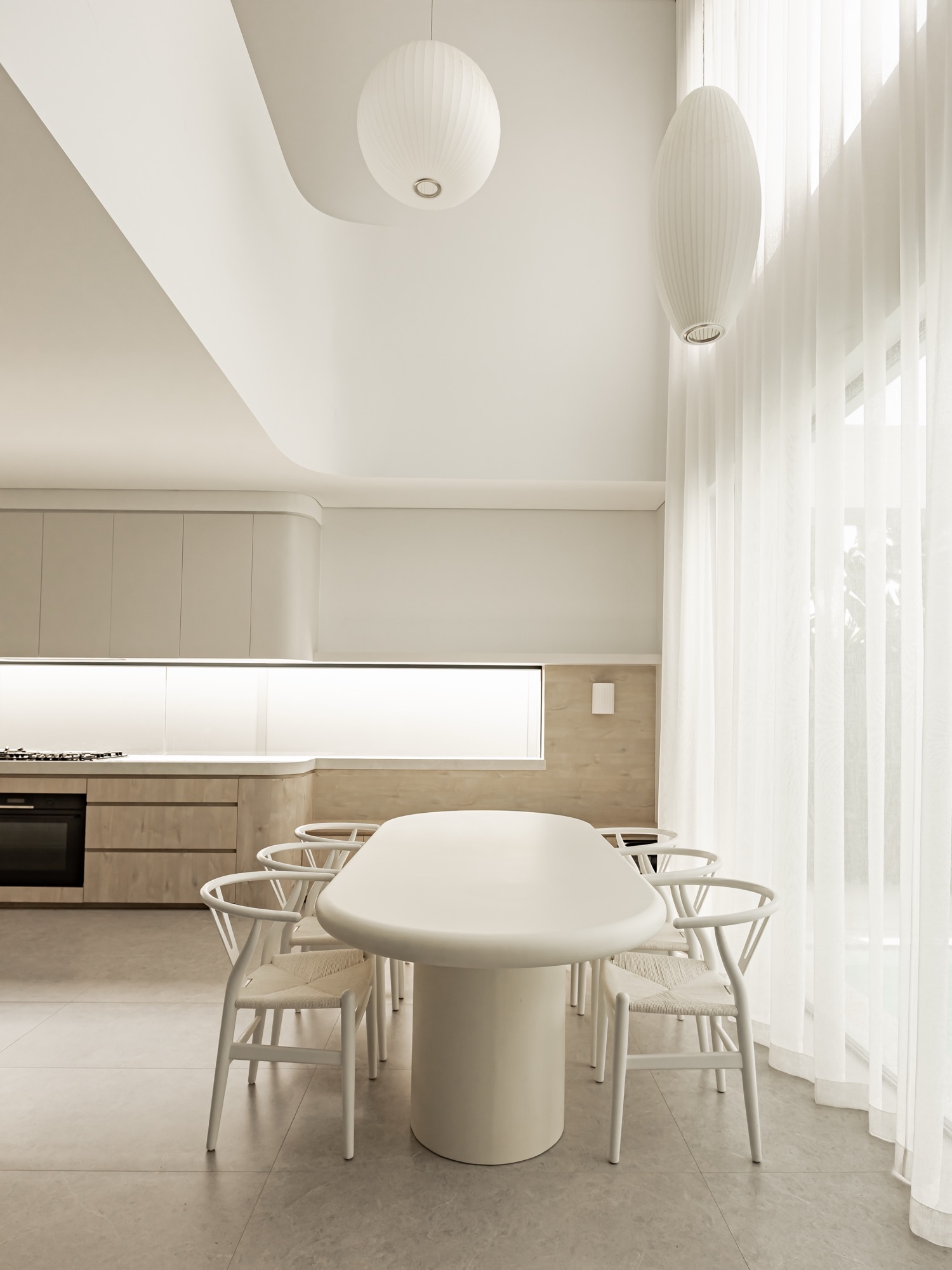 Dining table and woven chairs under paper pendants beside sheer curtains