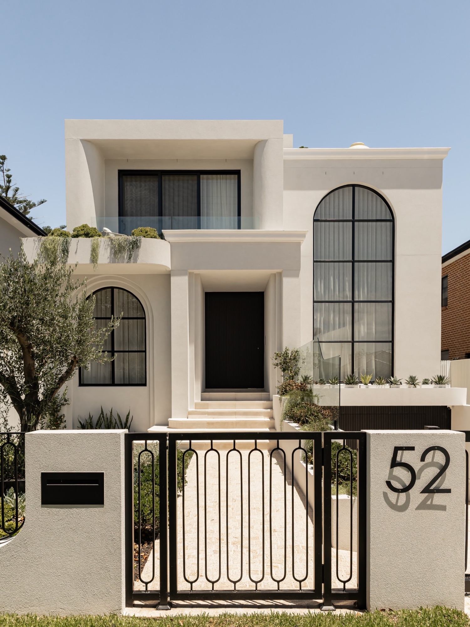 Rendered two-storey facade with arched black steel windows, olive tree and street gate